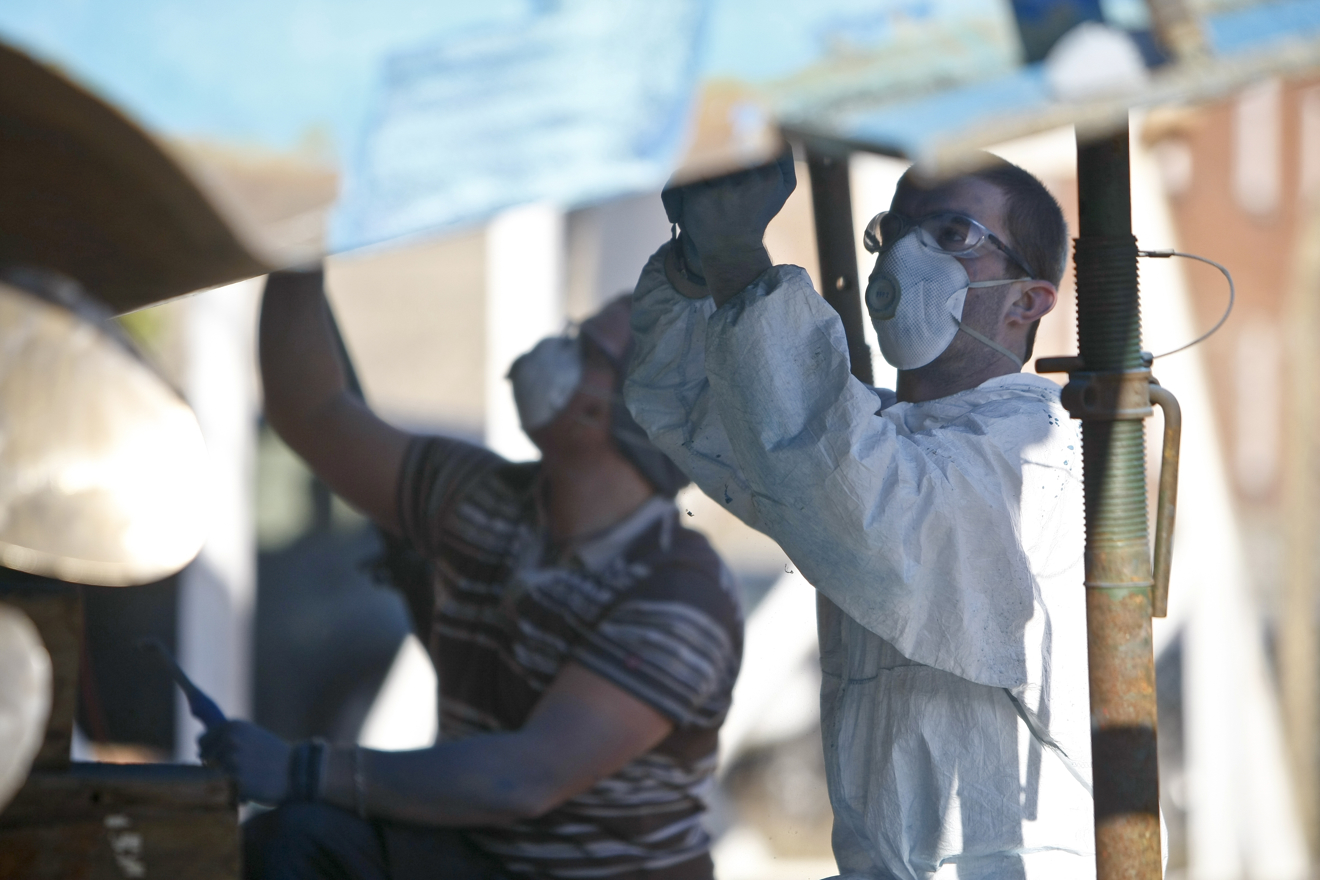 two men, one in protective clothing, both wearing masks underneath a yacht applying antifouling paint