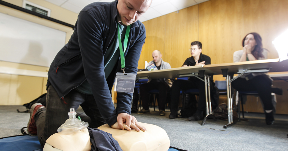Numerous people in a classroom at a First aid course, one man in knelt on the floor demonstrating CPR on a dummy